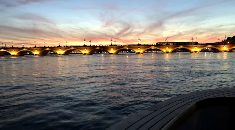 Vue du Pont de Pierre à Bordeaux au coucher de soleil avec des lumières illuminant les arches et le reflet sur la Garonne.